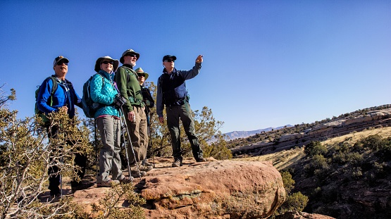 A Small Group of Mature Caucasian Men and Women Hike Together in the Rocky High Desert Mountains of Western Colorado on a Clear, Sunny Day and Stop on a Mountain Cliff to Point and Take in the View