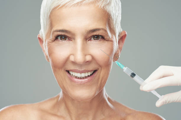 Beautiful smiling Caucasian senior woman standing in front of gray background and taking a shot of anti age product. Beauty photography. Beautiful smiling Caucasian senior woman standing in front of gray background and taking a shot of anti age product. Beauty photography. medical injection photos stock pictures, royalty-free photos & images