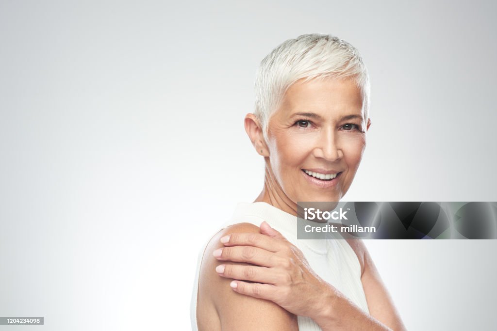 Schöne lächelnde Seniorin mit kurzen grauen Haaren posiert vor grauem Hintergrund. Schönheit Fotografie. - Lizenzfrei Frauen über 40 Stock-Foto