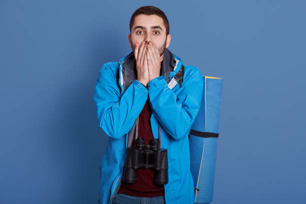istock Image of impressed confused handsome explorer covering his mouth with both hands, looking directly at camera, wearing blue jacket, dark blue sweatshirt, having all equipment for hiking alone. 1204238525