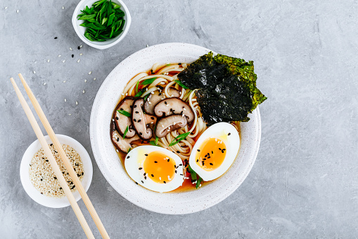 Asian Vegetarian Udon or Ramen noodles soup in bowl with Shiitake mushrooms, boiled eggs and nori sheets on grey stone background. Top view, copy space.