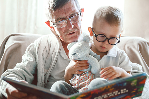 Elderly grandfather and little grandson read a book together and have fun sitting in a chair