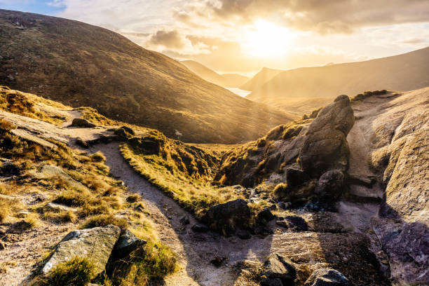 Footpath and large boulder leading to valley with water reservoir, dramatic sunset with sunrays. Mourne Mountains stock photo