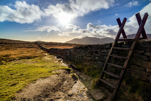 Mourn Wall with ladder on the bank of Slieve Donard mountain with blue sky, white clouds and sunrays. Mourne Mountains range in Northern Ireland