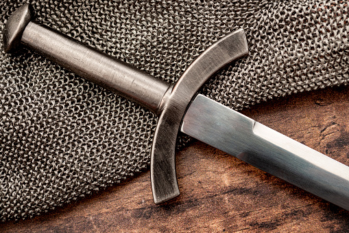 Medieval weapons and middle ages warfare conceptual idea with close up on sword on steel chain mail or maille on wooden background