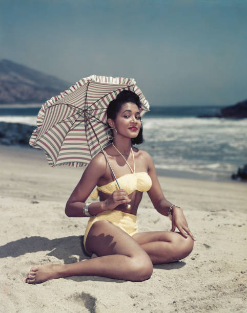 donna in bikini seduto sulla spiaggia con un ombrellone, sorridente, porta - archival foto e immagini stock