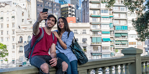 Panoramic photo of a young man and a young woman embracing and taking a selfie while sitting on a fence in Buenos Aires.
