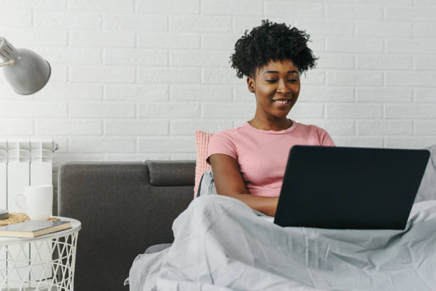 jeune femme afro-américaine de sourire utilisant l’ordinateur portatif dans le bâti le matin - using computer bedroom one woman only cheerful photos et images de collection