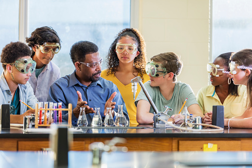 A mature African-American man in his 50s teaching a multi-ethnic group of teenagers in a high school science class. He is explaining a chemistry experiment with beakers and test tubes as the students look at him and listen intently.