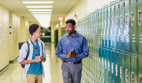 An African-American high school student walking with his teacher or school principal, in the hallway by a row of lockers. They are talking. The teacher has a serious expression on his face, a mature man in his 50s. The male student is an 18 year old teenage boy carrying a backpack.