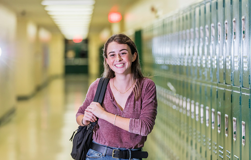 A high school student standing in the hallway of her school by a row of lockers. She is a teenage girl, 16 years old, carrying a book bag, looking at the camera, smiling.