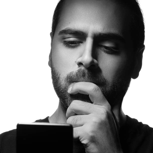 istock Arab guy using a smartphone and thinking. Black and white head shot of bearded Arab guy thinking while using phone 1204279353