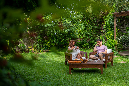 Young hispanic couple sitting in armchairs in the backyard of a house and talking while having coffee and yerba mate.