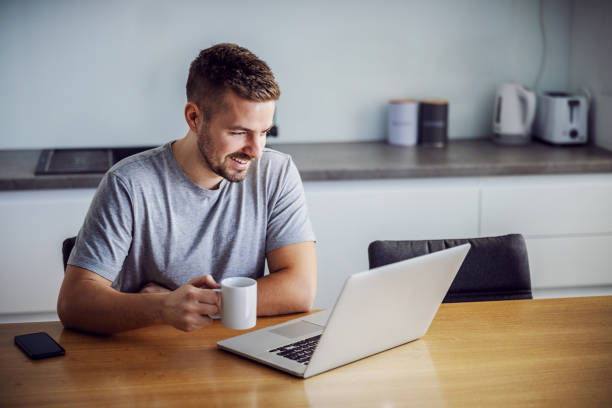 istock Young smiling man dressed casual sitting at dining table, holding mug with morning coffee and looking at laptop. He is visiting sites for online dating. 1204280977