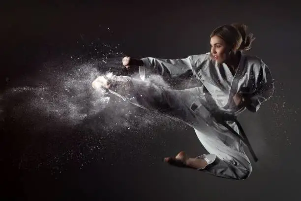 Energetic strong young karate girl bounces and makes a kick and hand smashing dust on a gray background. The concept of energy power and strength