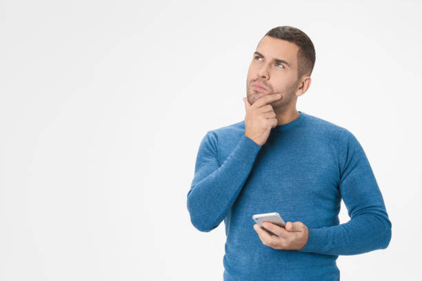 Handsome young man thinking while holding smartphone isolated over white background