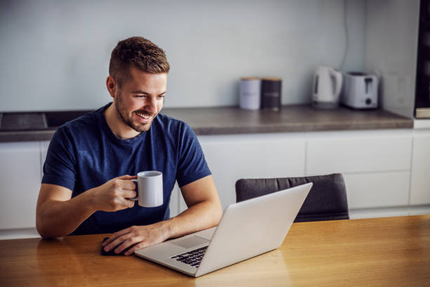 istock Young cheerful smiling man sitting at dinging table, holding mug with fresh morning coffee and looking at laptop. He is receiving likes for post on social media. 1204282288