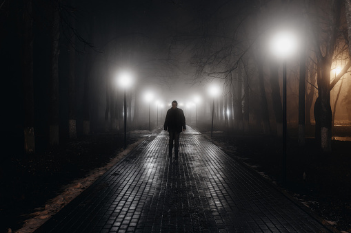 Sad man alone walking along the alley in night foggy park. Front view
