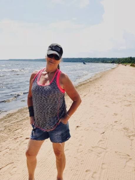 Fit senior woman with on Lake Michigan shoreline. Fit senior woman standing up proudly looking at the camera with USA hat and sunglasses on while enjoying The Lake Michigan shoreline in Gladstone, Michigan in The Upper Peninsula in the summer. gladstone michigan stock pictures, royalty-free photos & images