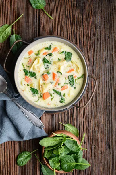 Comforting creamy Tortellini pasta soup with spinach, celery, carrot and chicken broth on rustic wooden background