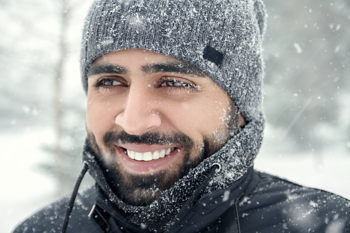 Happy bearded Middle eastern man wearing hat and winter clothes under the snow