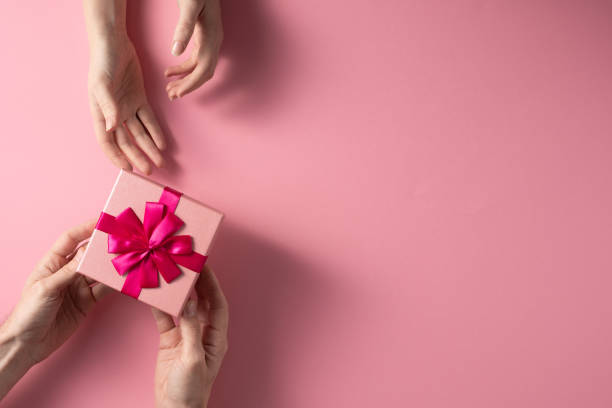 Valentine's Day celebration concept. A nice gift from a loved one. Box with a bow hands of a man and a woman on a delicate pink background. Copy space. Flat lay. stock photo