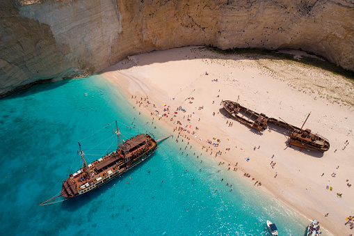 Lot of people on famous Navagio shipwreck beach on island of Zakynthos, Greece. One of the most popular beach on the world, recognizable by turquoise color of water and smuggler's ship that left as wreck. Tourist ship moored in shallow water. Aerial view photo made with drone.