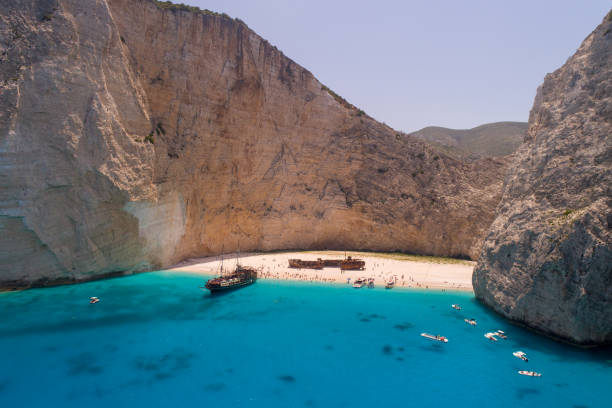 Lot of people on famous Navagio shipwreck beach on island of Zakynthos, Greece. One of the most popular beach on the world, recognizable by turquoise color of water and smuggler's ship that left as wreck. Tourist ship moored in shallow water. Aerial view photo made with drone.
