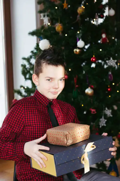 January 12, 2020 - Warsaw, Poland: Handsome teenage boy is sitting in front of a Christmas tree and holding his Christmas gifts on his knees before opening them