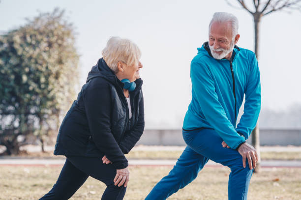 a senior adult male teaching a senior woman how to stretch in the park. the woman is wearing blue headset. - bending knees imagens e fotografias de stock