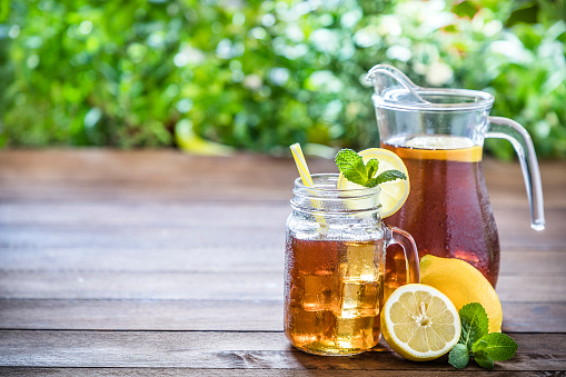 Front view of a drinking glass full of ice cubes and lemon ice tea alongside a whole and a sliced lemon, some mint leaves and a jug full of ice tea. The dinking glass and the jug are wet with condensed water and the glass has a yellow dinking straw, a lemon slice and a mint leaf on top. The objects are on a rustic wooden table and are at the right side of the image leaving a useful copy space at the left side. The main focus is on the drinking glass and the green background is defocused giving a feeling of freshness. Low key DSLR photo taken with Canon EOS 6D Mark II and Canon EF 24-105 mm f/4L