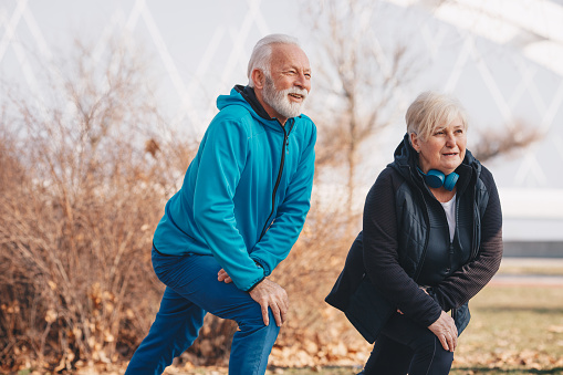An elderly man and an elderly woman stretching in the park. The woman is wearing headphones.