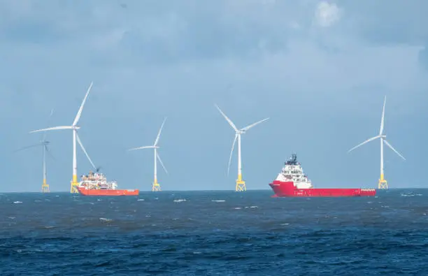 A group of tall offshore wind turbines in the North Sea, with cargo ships alongside them. These energy producing turbines are located off coastline of Aberdeen, on the east coast of Scotland. The renewable energy producers are an alternative to fossil fuel production, in an attempt to reduce the long term impact on our environment. They show a changing seascape along certain areas of the UK coastline, with shipping having to navigate around the manmade structures.
