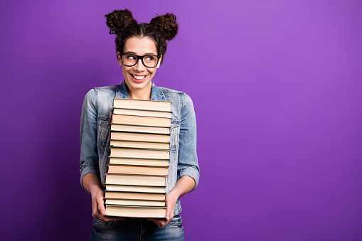 Photo of funny student lady holding many books carry literature home tricky, looking on librarian wear specs casual denim outfit isolated purple color background