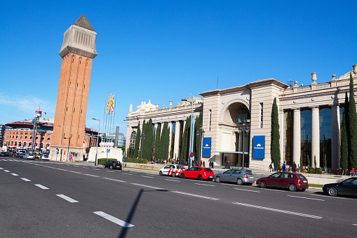Avinguda de la Reina Maria Cristina in Barcelona in winter with one tower of Torres Venecianes and at right side are Palau de Congressos de Catalunya and Placa de L'Univers. Cars are parked in street. People are on sidewalk. In left background is mall Arenas de Barcelona.