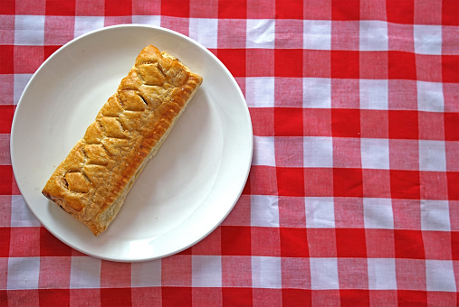 Flat lay of sausage roll on a plain white side plate on a red checked background with white space