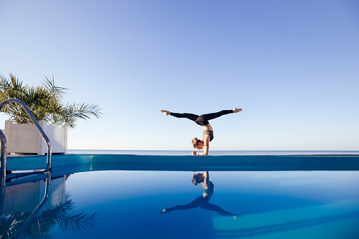 Young attractive woman practicing yoga stand in variation of Pincha Mayurasana exercise, handstand pose, working out by the pool, above the beach, relaxing against blue sky. Health and beauty concept
