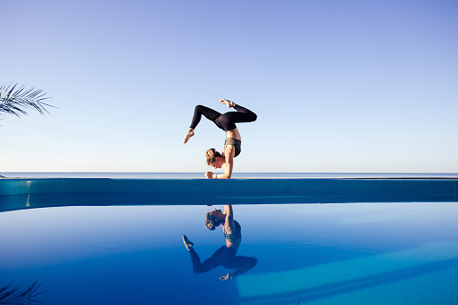 Young attractive woman practicing yoga stand in Adho Mukha Vrksasana exercise, Downward facing Tree pose, working out by the pool, above the beach, relaxing against blue sky. Health and beauty concept