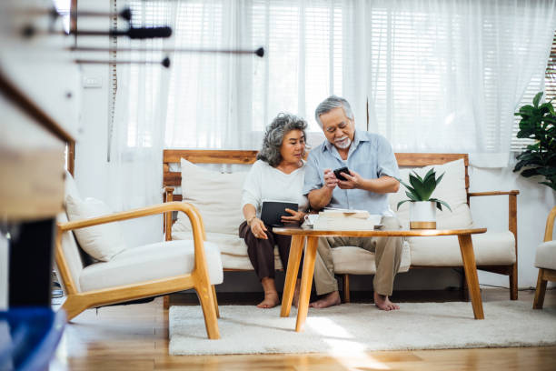 szczęśliwa azjatycka para seniorów za pomocą smartfona razem na kanapie w salonie, starszy mężczyzna sms-y wiadomości lub rozmowy z rodziną, starsza kobieta oglądania zainteresowanych gadżet , aging koncepcji społeczeństwa - grandparent family reading inside of zdjęcia i obrazy z banku zdjęć