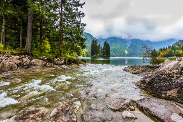 Waterfall from the lake Di Predil. Cold autumn in the Alps. Small island in the middle of the lake is overgrown with pine trees. The concept of active, mountain, environmental and photo tourism