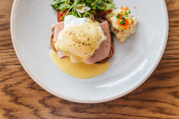 istock Top view of Eggs Benedict with ham, toast and mashed potato. Served with salad in white plate on wooden table. 1204450840