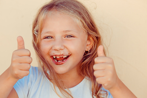 Yummy, I like it. The little girl who just ate a cake dirty of chocolate showing thumbs up gesture with hands, curly blonde tanned kid child. Funny picture