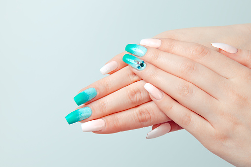 Art manicure. Trendy nail polish. Beautiful stylish woman hands with green white gradient pink pastel colors manicure with crystal gems isolated light blue background.