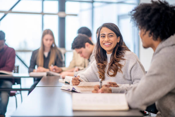 University Students in Class stock photo A group of multi-ethnic post secondary students sit in class with their textbooks open in front of them.  They are each individually studying.  The focus is on a girl of Indian decent who is looking up at her African peer and smiling. iberian ethnicity stock pictures, royalty-free photos & images