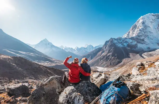 Photo of Young couple rejoicing on Everest Base Camp trekking route near Dughla 4620m. Backpackers left Backpacks and trekking poles and enjoying valley view with Ama Dablam 6812m peak and Tobuche 6495m