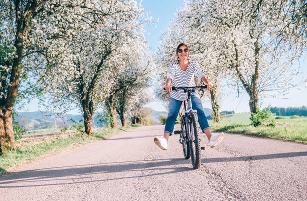 幸せな笑顔の女性は、リンゴの花の木の下の田舎道で自転車に乗ります - flower tree spring apple blossom ストックフォトと画像