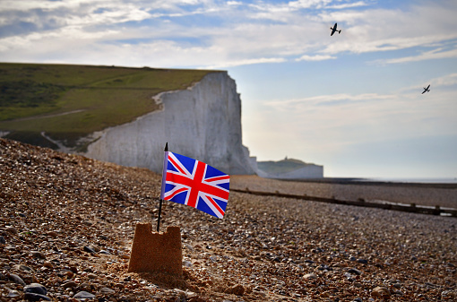 A sandcastle on an English Channel beach flying a British Union Jack flag beneath symbolic White Cliffs - a British metaphor. Overhead two legendary vintage World War II aircraft perform an aerobatic salute