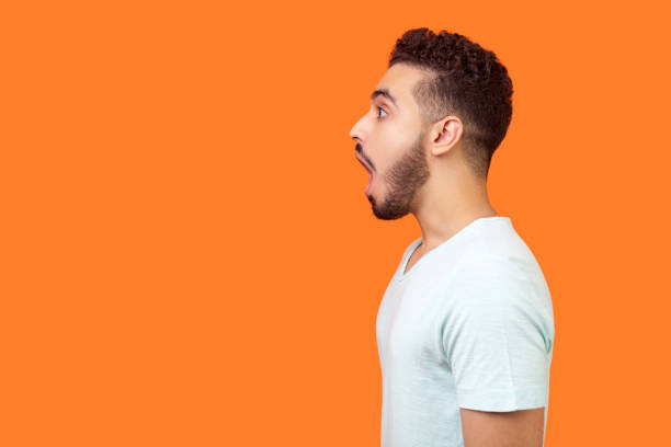 side view portrait of surprised brunette man looking left with big eyes and open mouth. studio shot isolated on orange background - wall profile imagens e fotografias de stock