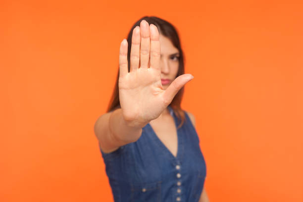 Prohibited! Strict brunette girl with serious bossy expression raising hand and showing stop Prohibited! Strict brunette girl with serious bossy expression raising hand and showing stop, caution, warning with raised palm, rejection gesture. indoor studio shot isolated on orange background stop gesture photos stock pictures, royalty-free photos & images