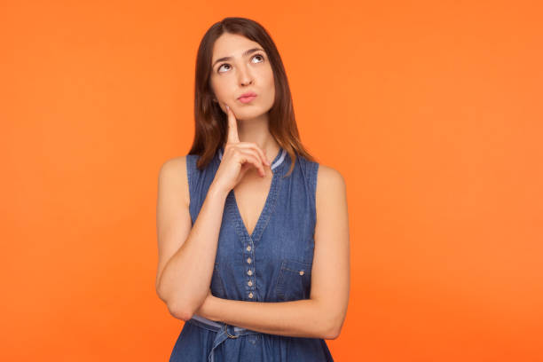 Pensive brunette woman in denim dress looking up with thoughtful doubtful expression Pensive brunette woman in denim dress looking up with thoughtful doubtful expression, pondering serious difficult idea, imagination and vision in mind. indoor studio shot isolated on orange background uncertainty stock pictures, royalty-free photos & images